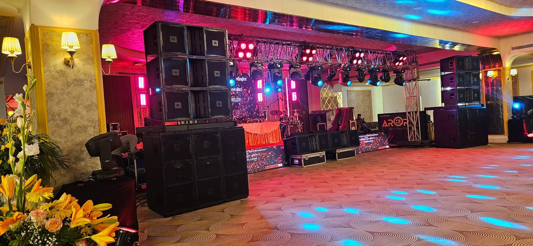 A banquet hall set for a function, with a stage backdrop, stacked speaker arrays and coloured stage lighting